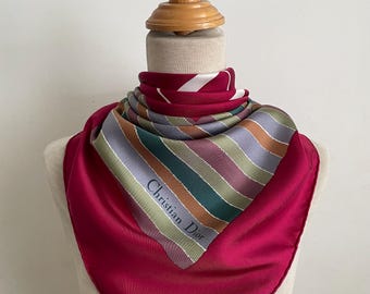 May include: A square silk scarf featuring a solid deep magenta border and a central panel with diagonal stripes in shades of green, brown, lavender, and white. The text Christian Dior is printed in a dark font on one of the striped sections.