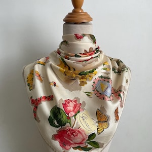 May include: A cream-colored silk scarf featuring a floral print with pink roses, yellow flowers, and various butterflies. The design includes a small illustration of a sailboat and the Gucci brand name printed in black script near a butterfly.
