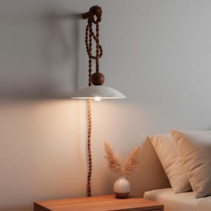 May include: A wall-mounted, minimalist pendant lamp with a white, disc-shaped shade. The lamp hangs from a brown rope and wooden accents. The lamp is above a wooden nightstand with a small vase of dried flowers.