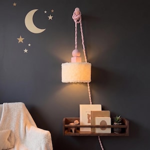 May include: A wall-mounted lamp with a fluffy, off-white shade and a pink rope cord. The lamp is suspended from a pink wooden fixture. A dark wooden shelf below holds books and a small plant. Gold stars and a crescent moon decorate the dark gray wall.