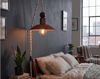 May include: A rustic, wall-mounted pendant lamp with a distressed, reddish-brown metal shade and a visible Edison-style bulb. The lamp hangs from a twisted rope cord and a wooden support. The bedroom setting includes a bed with neutral-colored bedding and a nightstand.