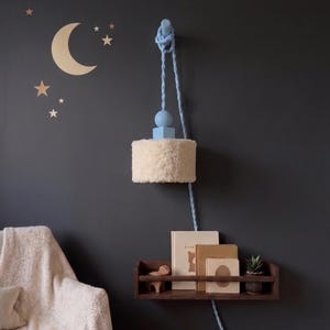 May include: A wall-mounted lamp with a fluffy, cream-colored shade and a light blue rope. The lamp hangs above a dark wooden shelf holding books and a small plant. The wall is dark gray, with gold star and moon decals.