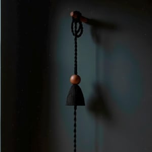 May include: A hanging black bell-shaped decoration with a wooden bead and a twisted black cord. The bell is suspended from a wooden dowel. The background is a dark teal color.