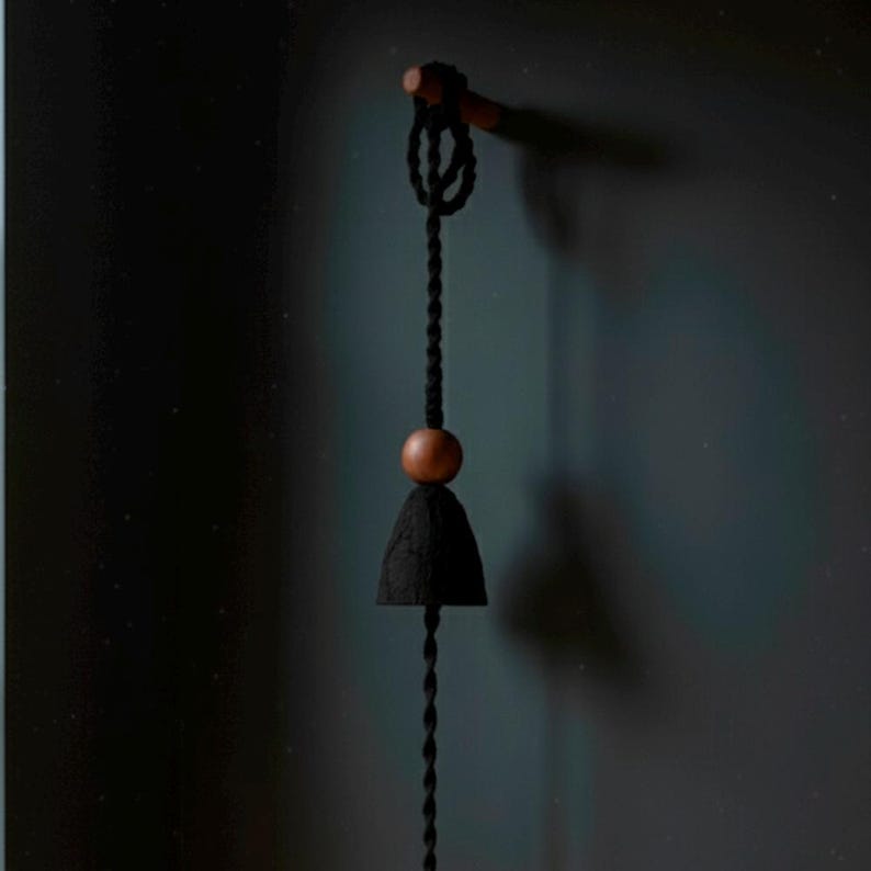 May include: A hanging black bell-shaped decoration with a wooden bead and a twisted black cord. The bell is suspended from a wooden dowel. The background is a dark teal color.