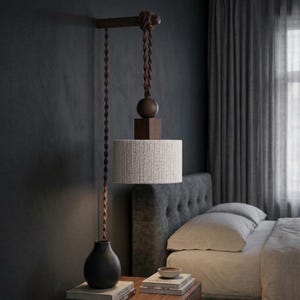 May include: A modern pendant light fixture with a cylindrical lampshade wrapped in white fabric. The light hangs from a wooden beam and features a dark brown twisted cord. A dark vase sits below the light, on a wooden nightstand.