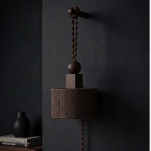 May include: A brown wall-mounted lamp with a cylindrical shade made of tightly wound rope. The lamp features a square base, a spherical accent, and a twisted cord. The fixture is mounted on a dark wall, creating a minimalist aesthetic.