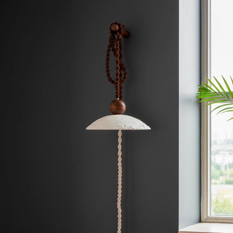 May include: A hanging pendant light fixture with a white, textured shade and a long, cream-colored rope. The fixture is suspended from a dark brown, twisted rope and a wooden sphere. The fixture is mounted on a dark gray wall.