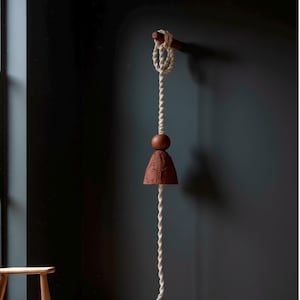 May include: A hanging decorative bell with a natural, earthy aesthetic. It features a wooden dowel, a knotted rope, a wooden sphere, and a textured, bell-shaped element in a reddish-brown hue. The bell hangs against a dark wall.