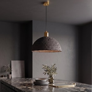 May include: A contemporary pendant light featuring a textured, dark grey lampshade and a gold-coloured ceiling fitting. The light is suspended by a black cable, suitable for a kitchen or dining space. The lampshade has a distinctive, rough texture, adding an industrial design element.