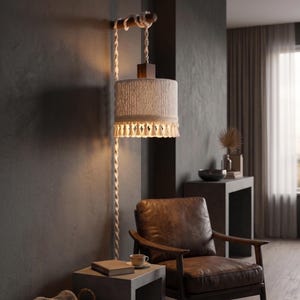May include: A wall-mounted lamp featuring a cylindrical shade crafted from off-white macrame cord with a fringed bottom edge. The shade hangs from a dark wooden horizontal arm by a twisted macrame cord that extends down the wall.