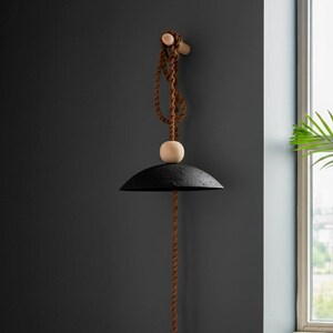 May include: A black, textured pendant lamp with a rounded shade, suspended by a brown rope and a wooden dowel. A small, light-colored wooden sphere is positioned between the shade and the rope. The lamp hangs against a dark gray wall.