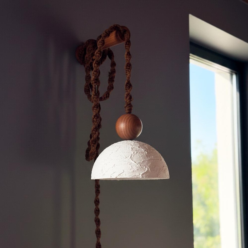 May include: A wall-mounted light fixture with a white, textured lampshade. The fixture features a wooden arm and a brown rope cord. A wooden sphere sits atop the lampshade. The fixture is mounted on a gray wall near a window.