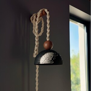 May include: A modern, handcrafted wall-mounted lamp. It features a textured black lampshade, a wooden sphere, and a cream-colored macrame rope. The lamp is attached to a wooden rod and hangs near a window.
