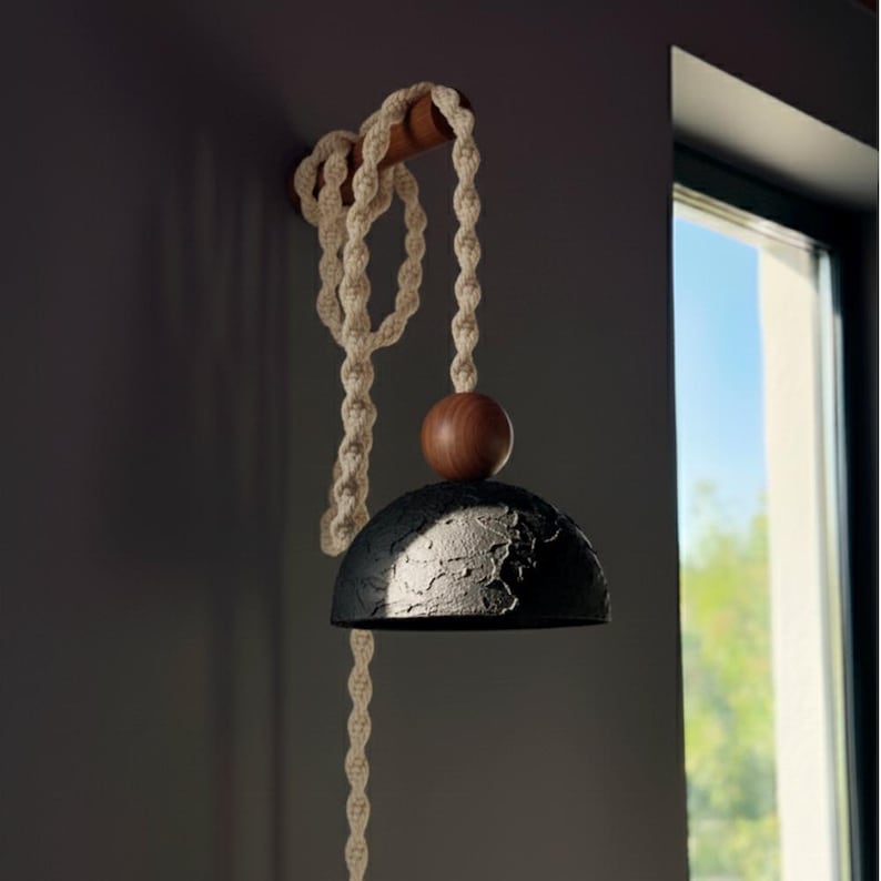 May include: A modern, handcrafted wall-mounted lamp. It features a textured black lampshade, a wooden sphere, and a cream-colored macrame rope. The lamp is attached to a wooden rod and hangs near a window.