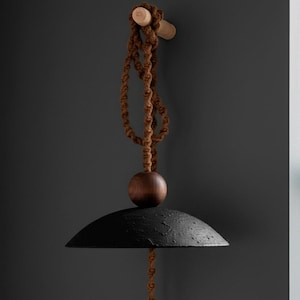 May include: A black, textured pendant light with a rounded shade, suspended by a brown rope and wooden accents. The light fixture is attached to a wooden peg on a dark gray wall. The design is minimalist and modern.