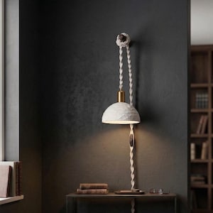 May include: A unique wall-mounted lamp with a textured white shade and a brass-colored accent. The lamp hangs from a twisted rope and is mounted on a dark wall. The lamp is turned on, illuminating the area below. A small table holds books and glasses.