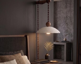 May include: A wall-mounted pendant light featuring a dark brown wooden horizontal support bar, a twisted dark brown cord, and a textured white dome-shaped shade. A small wooden sphere sits atop the shade, which emits a warm light downward.