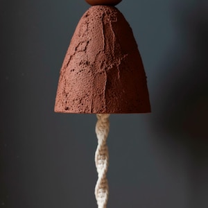 May include: A handcrafted, terracotta-colored pendant lamp with a textured, cone-shaped shade. The lamp features a twisted, off-white macrame cord and a small, round, brown accent at the top. The background is a muted gray.
