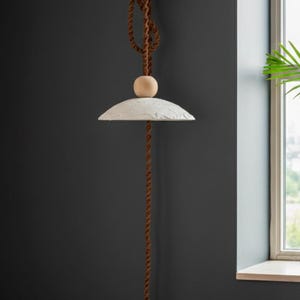 May include: A modern pendant light with a white, textured shade and a wooden sphere accent. The light hangs from a brown, twisted rope. The design is minimalist and suitable for various interior styles.