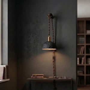 May include: A hanging pendant lamp with a dark, textured shade and a gold-colored top. The lamp is suspended by a twisted brown cord and is mounted on a dark wall. The lamp is casting a warm light.