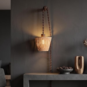 May include: A wall-mounted pendant light featuring a conical shade made of light-colored woven natural fibers. The lamp hangs from a dark brown twisted cord that loops over a circular wooden wall mount, emitting a warm glow from the center.