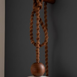 May include: Close-up of a brown macrame hanging light fixture. The fixture features a knotted rope design, a wooden sphere, and a wooden rod. The background is a dark gray wall.