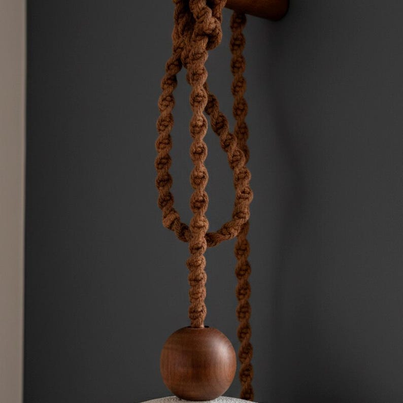 May include: Close-up of a brown macrame hanging light fixture. The fixture features a knotted rope design, a wooden sphere, and a wooden rod. The background is a dark gray wall.