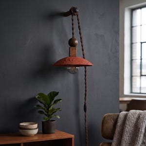Könnte beinhalten: Eine Wandlampe mit terrakottafarbenem Schirm und Holzsockel. Die Lampe hängt an einem braunen Seil und hat eine sichtbare Glühbirne. Das Design integriert natürliche Materialien und eine Vintage-Ästhetik, passend für die Inneneinrichtung.