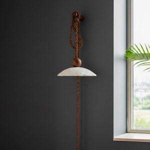 May include: A modern pendant light fixture with a textured, off-white shade. The light hangs from a brown rope and wooden ball, attached to a wooden fixture on the wall. The design is minimalist and rustic, suitable for interior decor.