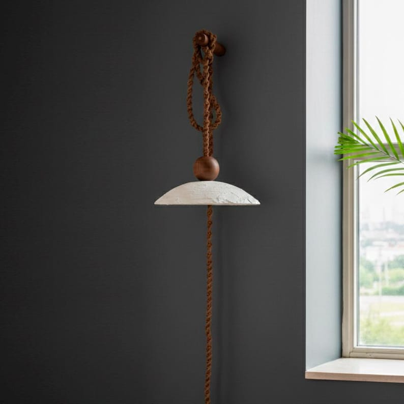 May include: A modern pendant light fixture with a textured, off-white shade. The light hangs from a brown rope and wooden ball, attached to a wooden fixture on the wall. The design is minimalist and rustic, suitable for interior decor.