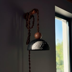 May include: A modern, handcrafted pendant light fixture. The fixture features a textured, dark gray dome-shaped shade, a wooden sphere, and a brown rope cord. The light is mounted on a wooden bracket, creating a warm, inviting ambiance.
