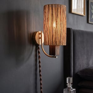 May include: A wall-mounted lamp with a cylindrical shade made of vertical, textured, light brown material. The lamp has a brass-colored arm and base, and a dark brown, twisted cord. The lamp is mounted on a dark gray wall.
