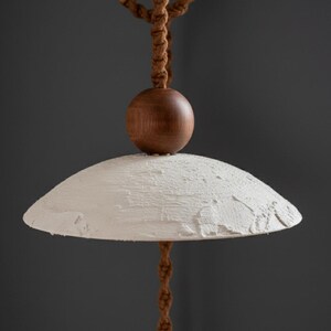 May include: A modern pendant light with a textured, off-white shade and a dark wooden sphere. The light hangs from a brown rope, creating a natural and minimalist aesthetic. The shade has a diameter of approximately 16 inches.