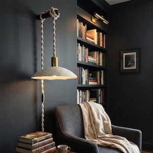 May include: A wall-mounted pendant light with a gray paper shade and a brass fixture. The light hangs from a wooden beam and rope. The room features a bookshelf, a gray armchair, and a stack of books.