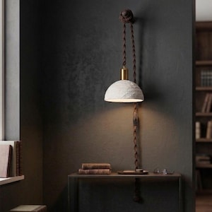 May include: A unique, textured white pendant lamp with a brass-colored top hangs from a dark brown twisted cord. The lamp is mounted on a dark gray wall, casting a warm glow. A small table below holds books and a tray.