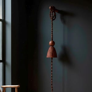 May include: A rustic, brown wind chime hanging on a dark wall. The chime features a twisted rope, a wooden sphere, and a bell-shaped element. The design is simple and elegant, suitable for indoor or outdoor decor.