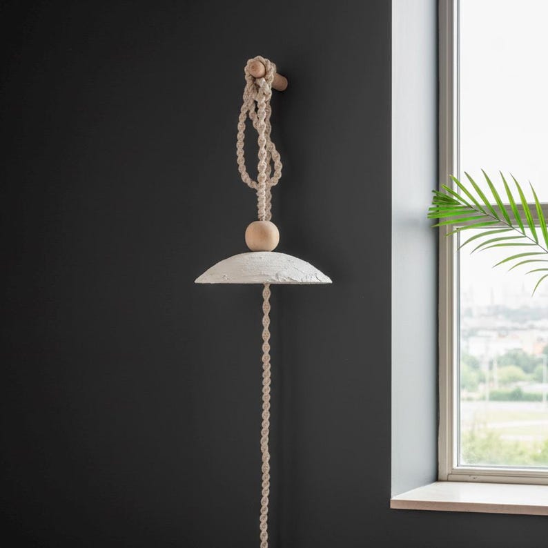May include: A hanging pendant light with a textured, off-white shade and a natural rope cord. The light is suspended from a wooden dowel and features a wooden bead. The design is minimalist and bohemian, suitable for interior decor.