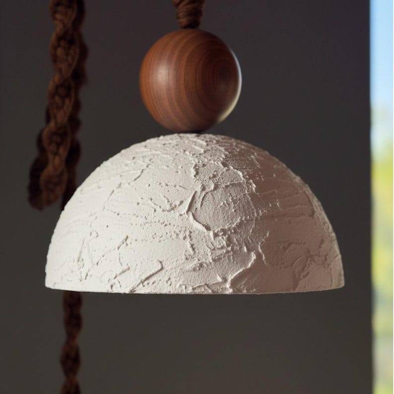 May include: A white, textured, semi-spherical pendant lamp with a wooden sphere on top. A brown macrame cord hangs from above. The lamp has a rustic, handmade aesthetic, suitable for home decor.