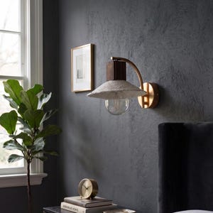 May include: A wall-mounted light fixture with a textured, off-white shade and a clear bulb. The fixture has a dark brown wooden block and a curved brass arm. The fixture is mounted on a dark gray wall. A gold-framed picture hangs on the wall.