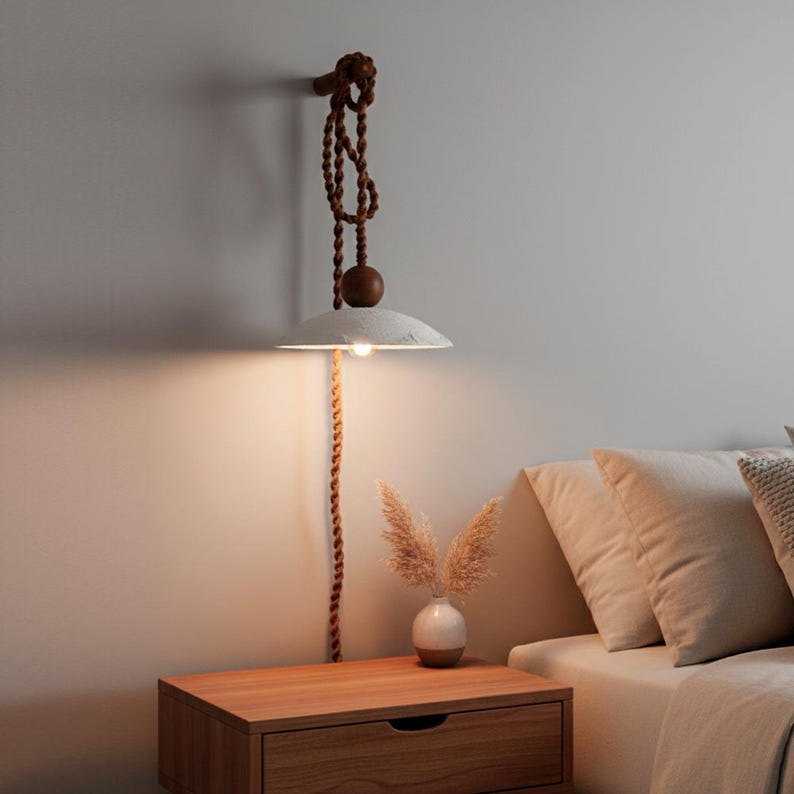 May include: A wall-mounted, minimalist pendant lamp with a white, disc-shaped shade. The lamp hangs from a brown rope and wooden accents. The lamp is above a wooden nightstand with a small vase of dried flowers.
