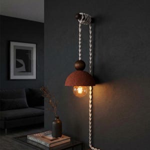 May include: A contemporary wall light with a red-brown lampshade and a warm bulb. The light is suspended by a white rope and has a wooden accent. The design is minimalist and stylish, suitable for interior decor. The light is mounted on a dark grey wall.