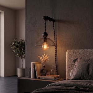 May include: A wall-mounted pendant light featuring a matte black fixture, a braided cord, and a clear glass dome shade. The light emits a warm glow from an exposed filament bulb, hanging above a bedside table with books and a decorative vase.