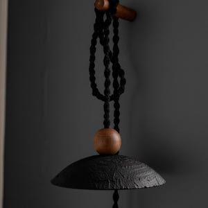 May include: A black, textured metal wind chime with a wooden bead and a wooden bar for hanging. The chime hangs from a black rope against a dark gray background. The bell-shaped chime has a rustic, handcrafted appearance.