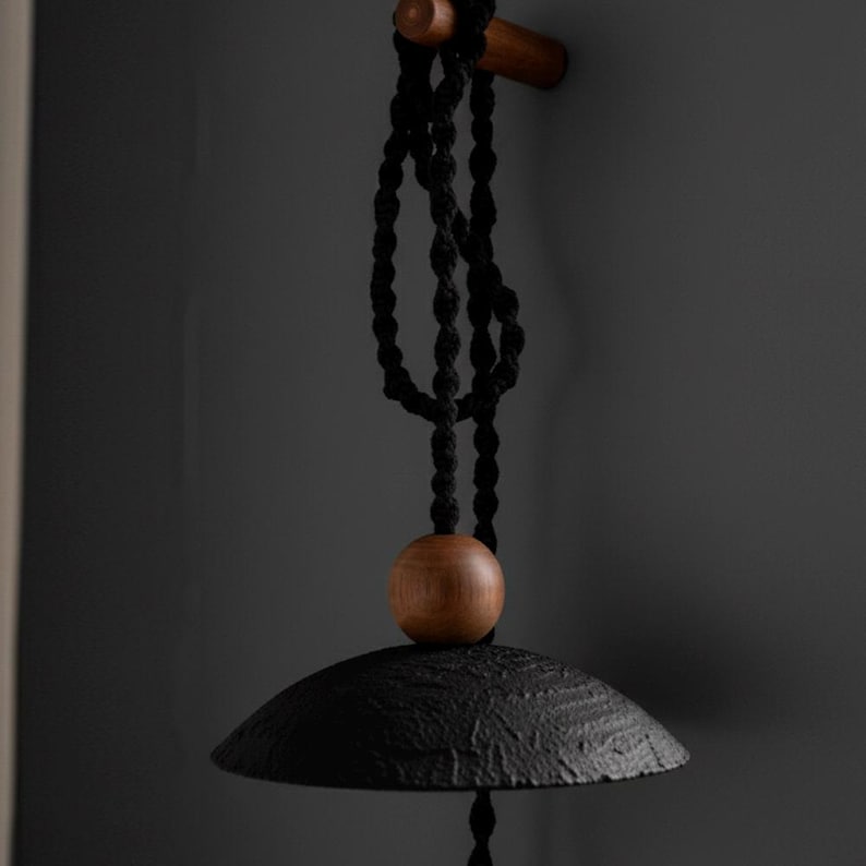 May include: A black, textured metal wind chime with a wooden bead and a wooden bar for hanging. The chime hangs from a black rope against a dark gray background. The bell-shaped chime has a rustic, handcrafted appearance.