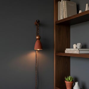 May include: A wall-mounted, bell-shaped lamp with a reddish-brown shade and a warm, inviting glow. The lamp is suspended by a twisted brown rope and features a wooden sphere. The design is minimalist and modern, suitable for interior decor.