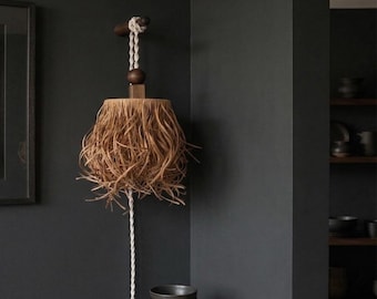 May include: A wall-mounted light fixture featuring a textured, light brown raffia lampshade suspended by a white twisted rope from a dark wooden peg. The fixture includes a dark wooden bead accent and a white cord extending downward.