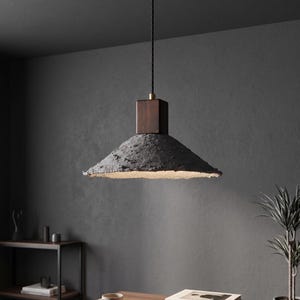 May include: A modern pendant light with a dark gray, textured shade and a square, dark wood top. The light hangs from a black cord and features a brass accent. The shade has a rough, uneven edge, and the interior is a warm, off-white color.