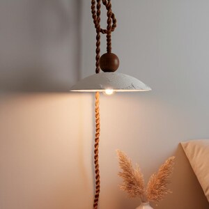 May include: A hanging pendant lamp with a textured, off-white shade and a warm light bulb. The lamp is suspended by a brown rope, with a wooden sphere accent. The design has a bohemian or rustic aesthetic, suitable for interior decor.