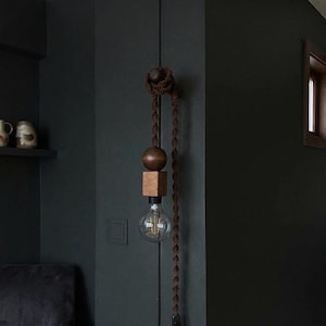 Può includere: Una lampada a sospensione decorativa con un design a sfera e cubo in legno, sospesa da una corda marrone. La lampadina è visibile sotto i componenti in legno. La lampada è montata su una parete scura, creando un'estetica minimalista.