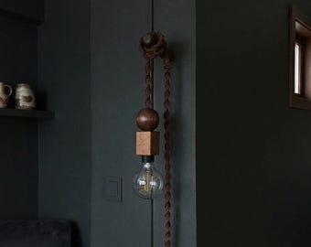 May include: A hanging light fixture with a wooden sphere and cube design, suspended by a brown rope. The light bulb is visible below the wooden components. The fixture is mounted on a dark wall, creating a minimalist aesthetic.